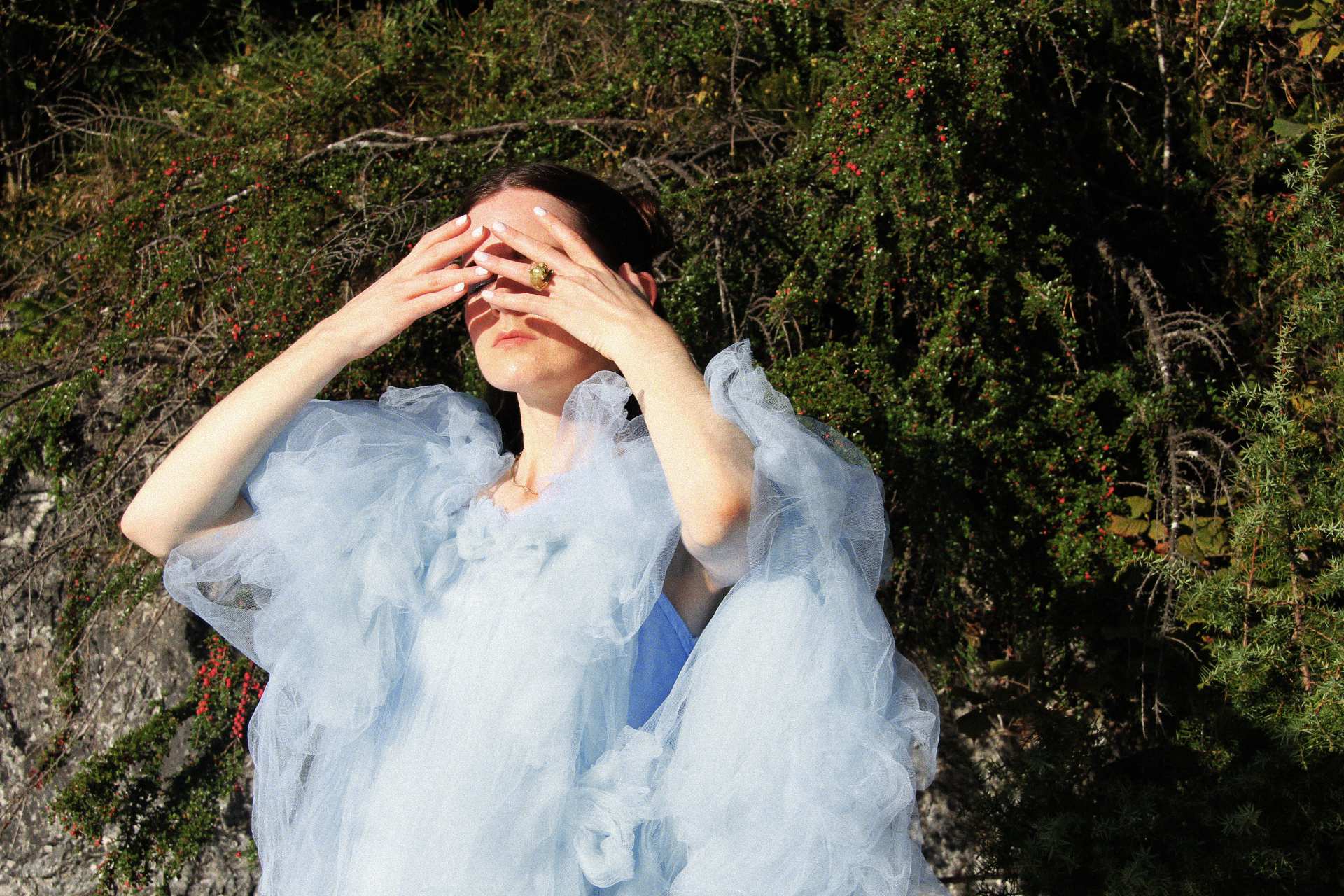 MOLTENO in pale blue tulle, hands over her eyes, in front of a berried hedge.