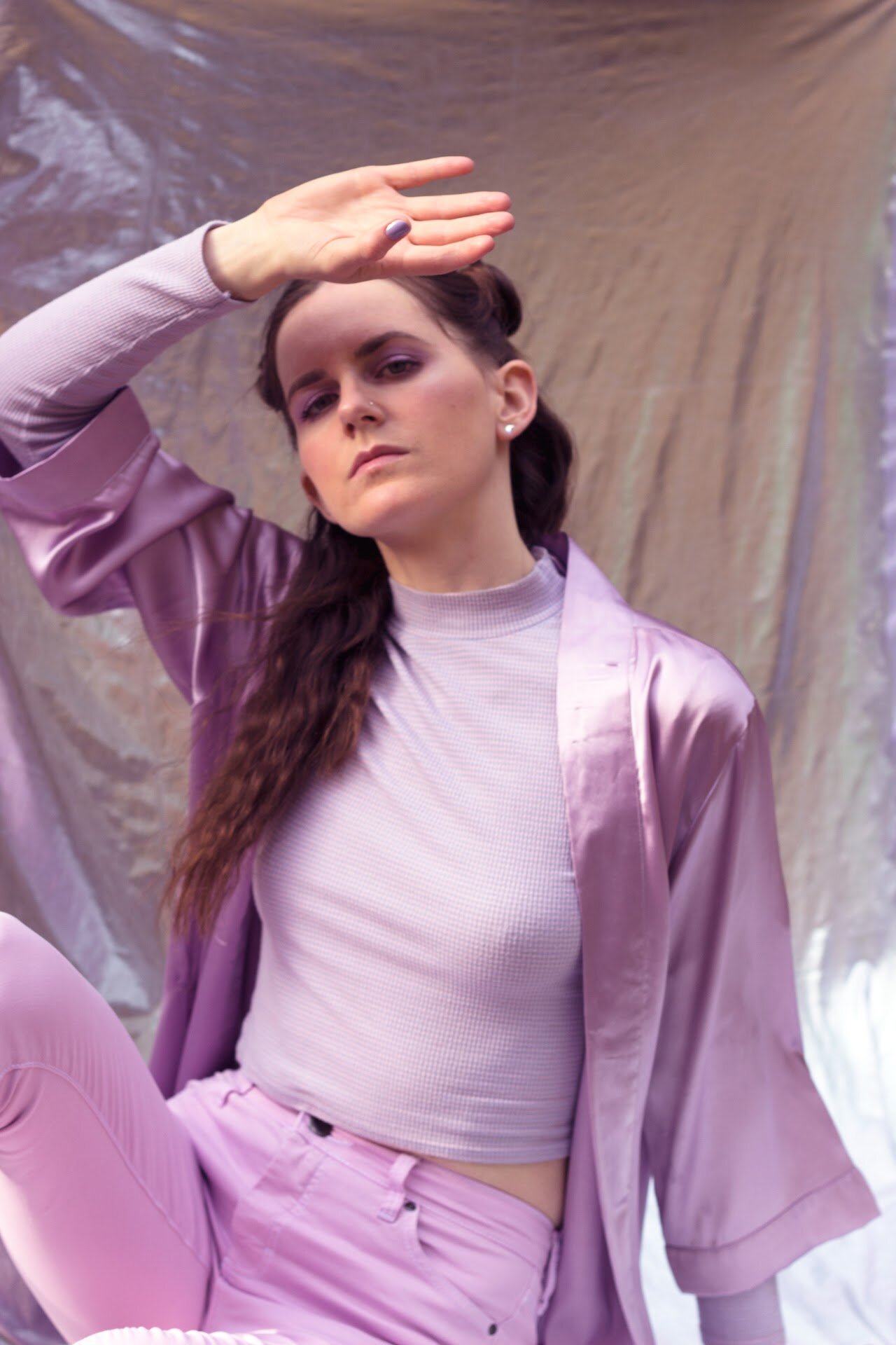 MOLTENO in a lilac satin shirt and lilac jeans, hand raised to her brow, in front of iridescent foil.