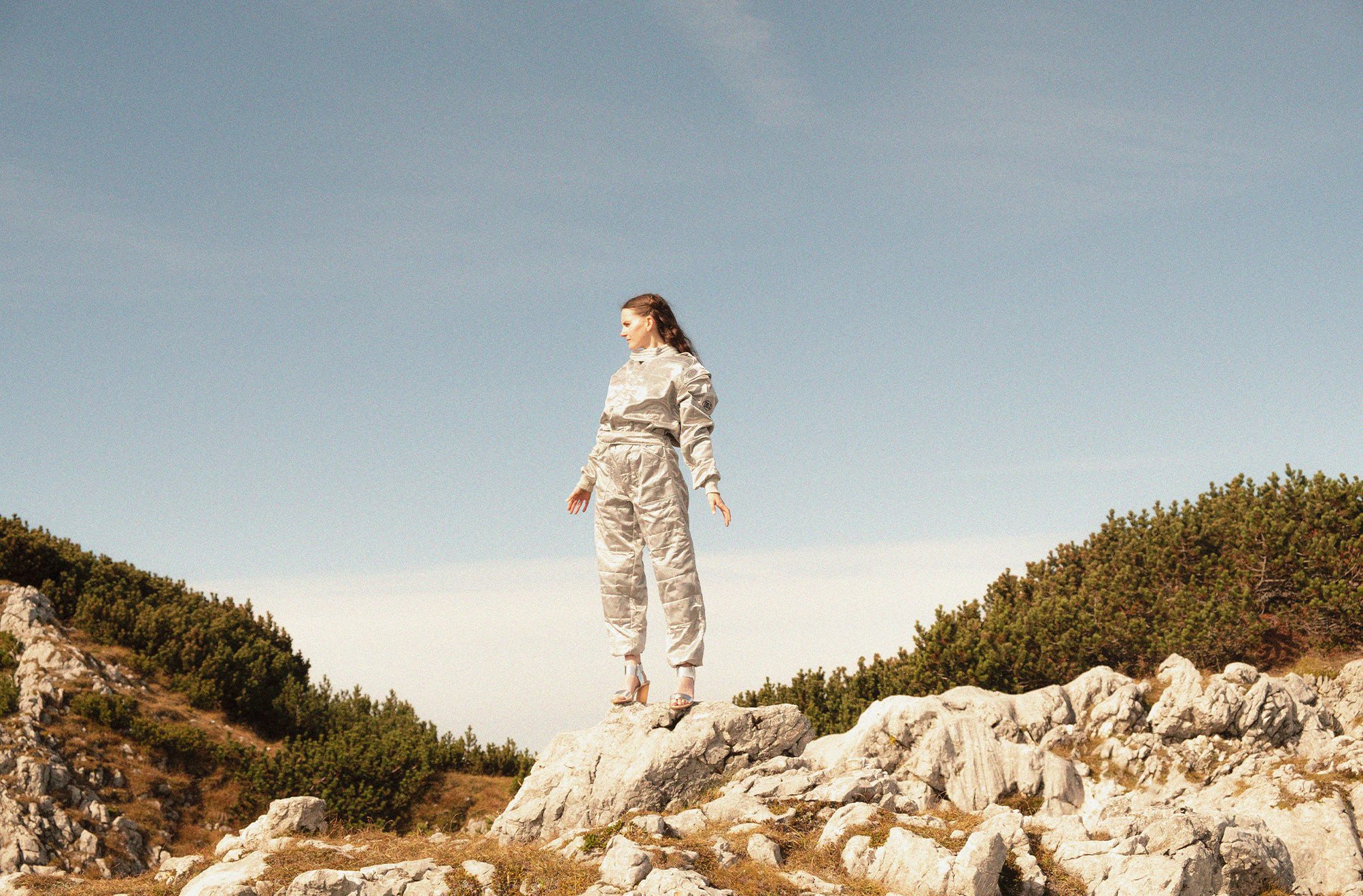 MOLTENO in a silver quilted suit standing on a rocky outcrop against a pale blue sky.