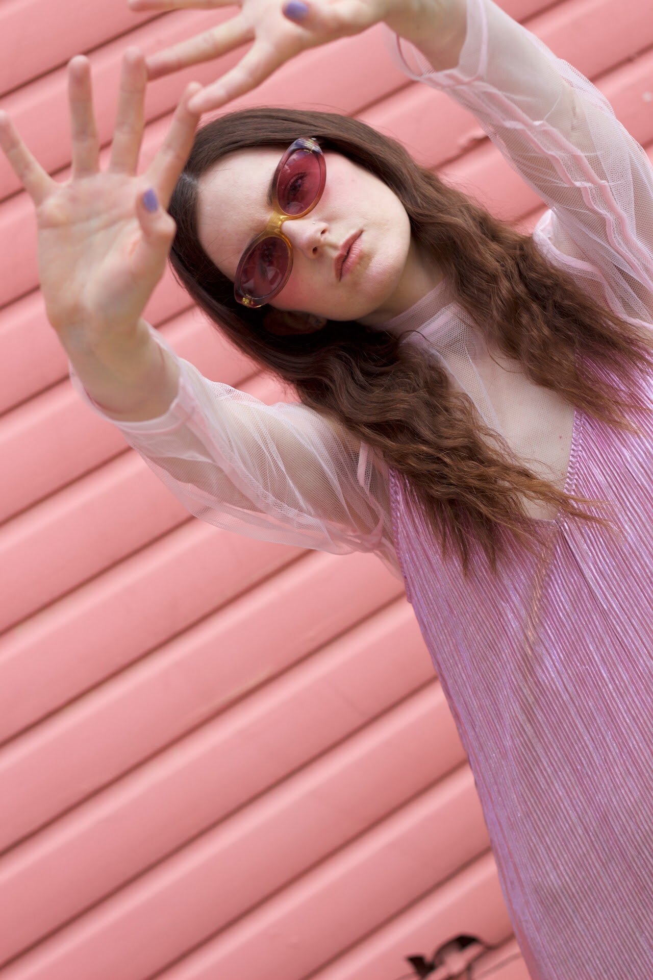 MOLTENO in pink tinted sunglasses and a pink pleated dress, hands raised, against a pink wall.