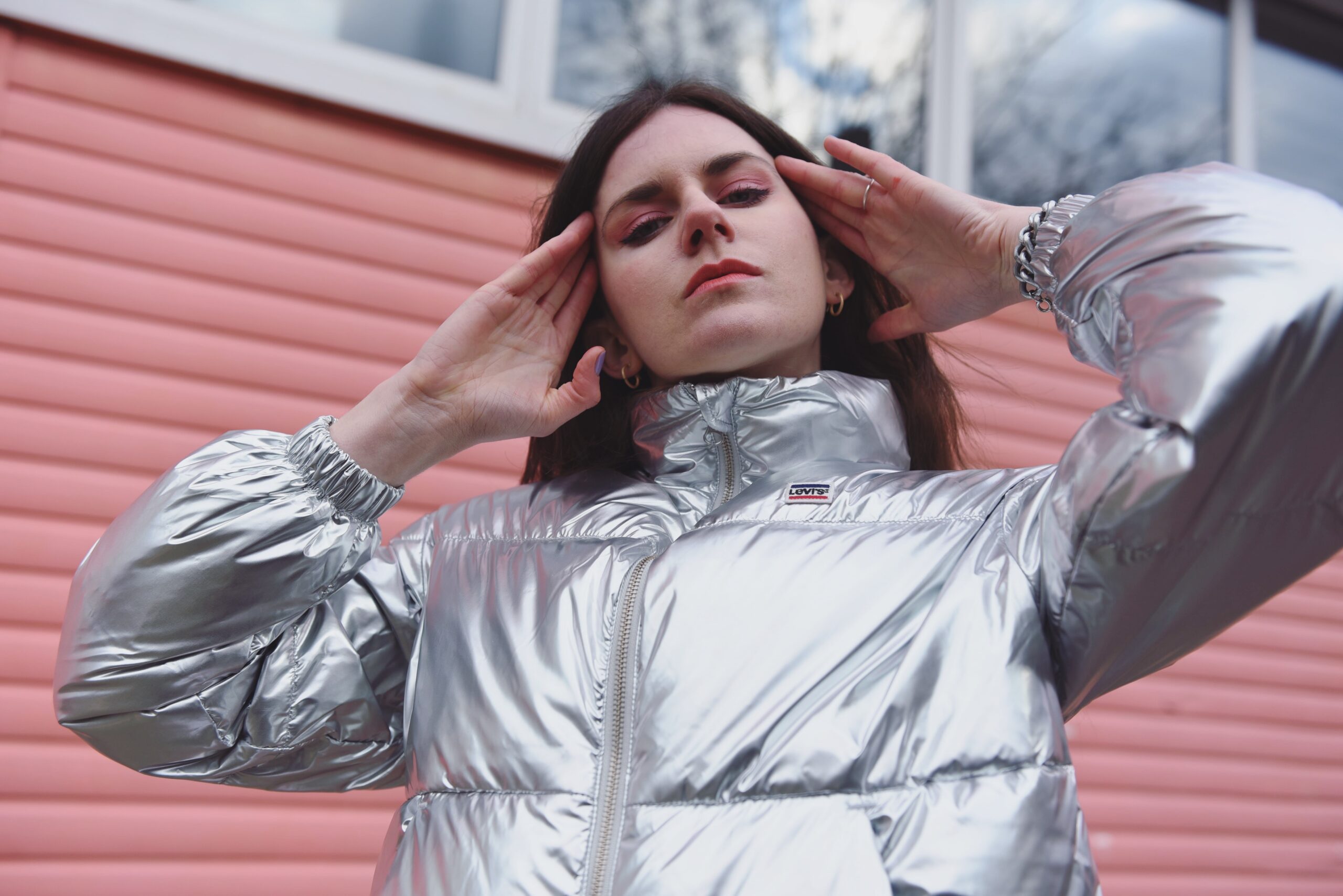 MOLTENO in a silver puffer jacket, hands at her temples, against a pink clapboard wall.