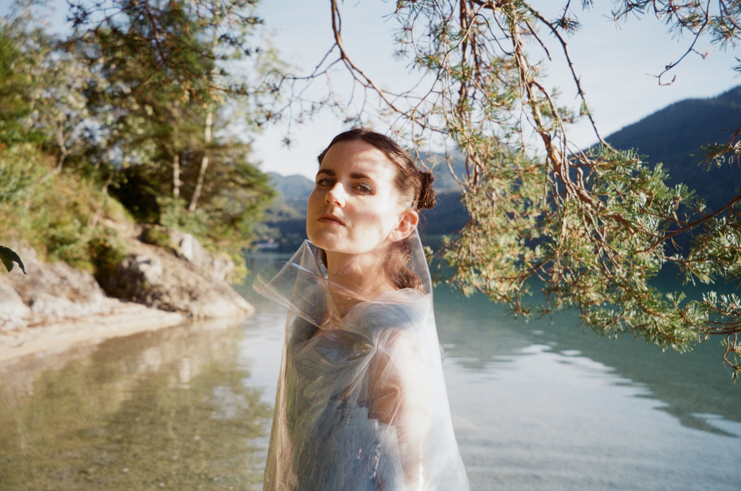 MOLTENO standing in a lake, wrapped in clear plastic and blue tulle, pine branches overhead.